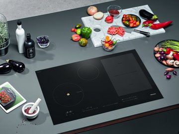 Induktionsherdplatte mit vielen Lebensmitteln drum herum plus Tablet mit Miele CookAssistant-App