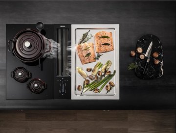 Edelstahl-Grillplatte auf einem Induktionsherd, darauf Lachs und Gemüse. Daneben steht ein Kochtopf mit Qualm der in eine Integrierte Downdraft-Haube gezogen wird.